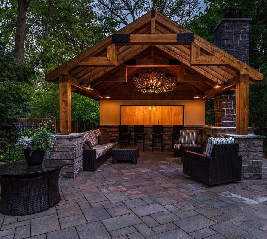 A cozy outdoor pavilion with wooden beams, a chandelier, wicker seating, and a bar area, surrounded by trees and lit warmly