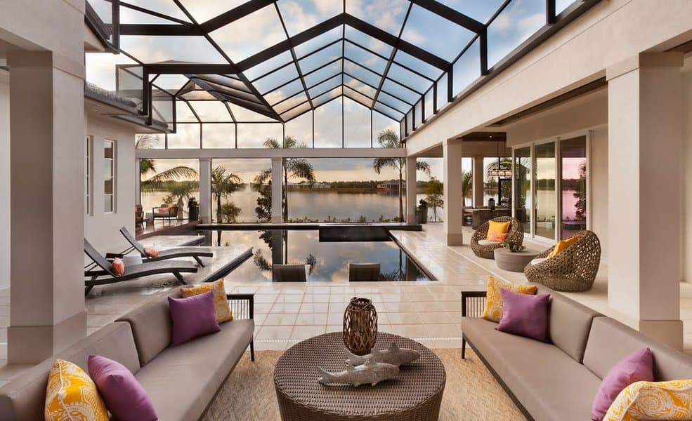 Modern patio with sofas, colorful pillows, a central table, and loungers by a pool; screened enclosure offers lake and sky views