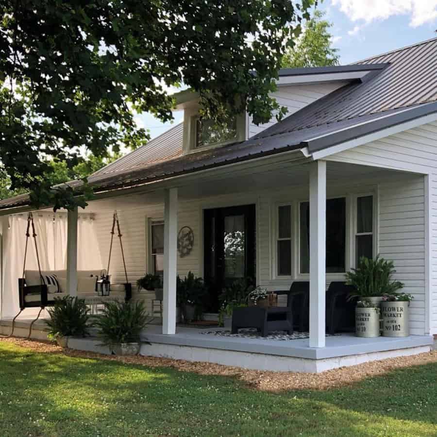 White house with a covered porch, swing chairs, and green plants