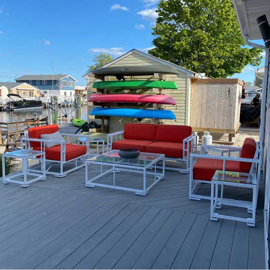 Outdoor deck with modern white metal furniture, red cushions, and glass tables; kayaks are stacked by a wooden shed; houses and a waterway are visible