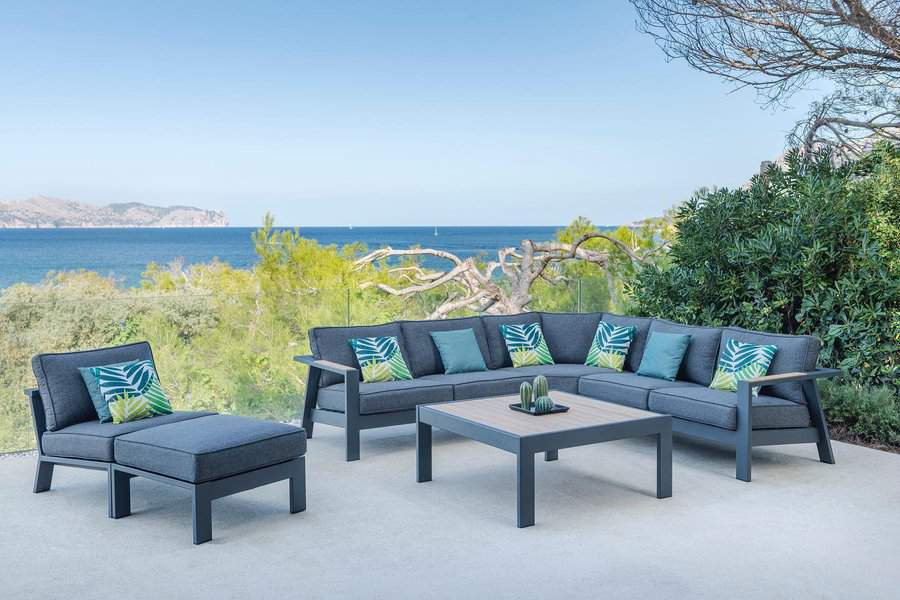 Outdoor patio set with blue cushions and tropical pillows, positioned near a seaside view with trees and clear sky in the background