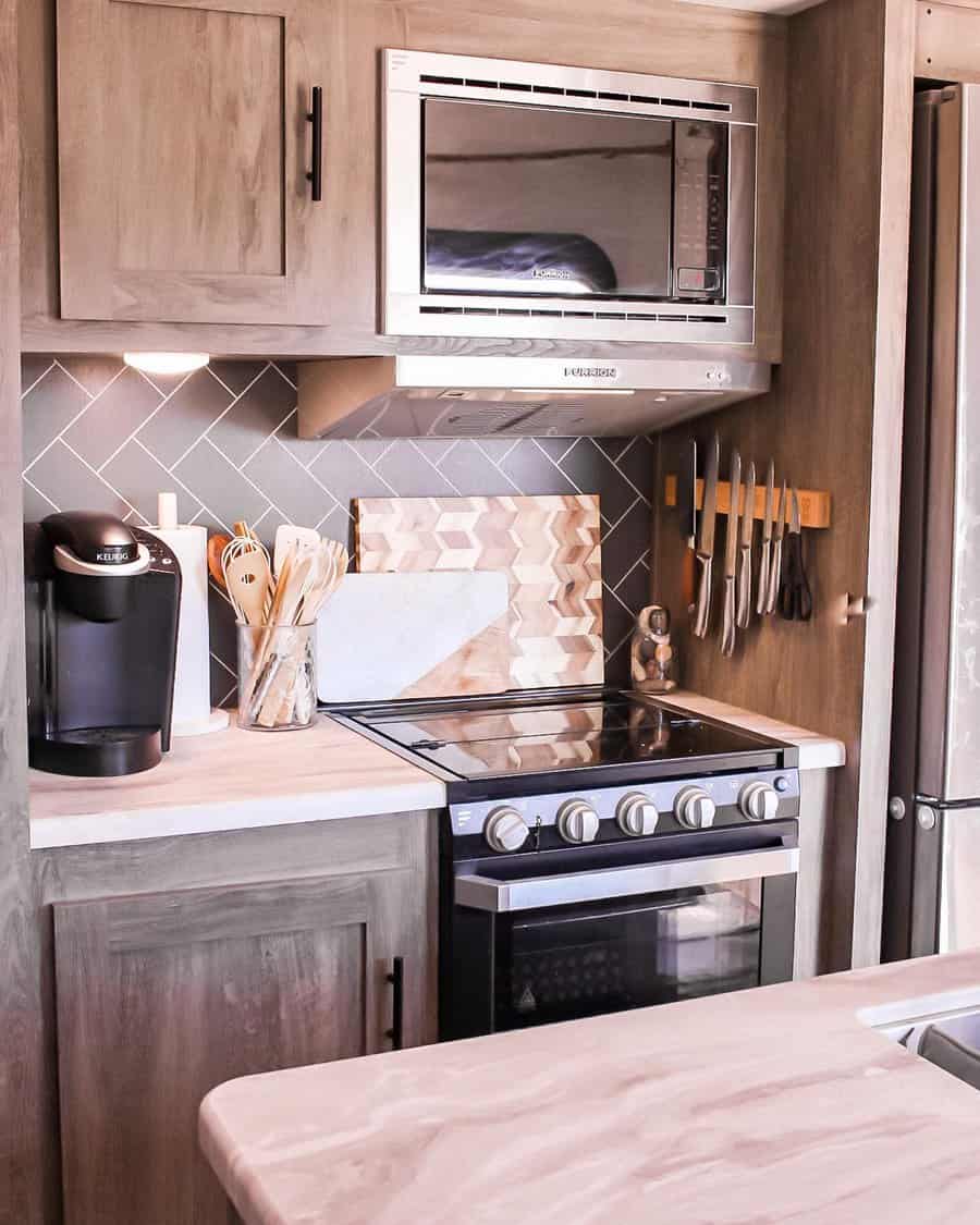 A sleek and modern RV kitchen featuring a stainless steel microwave, oven, and a coffee maker, with geometric tiles and neatly organized utensils, offering a functional and stylish space.