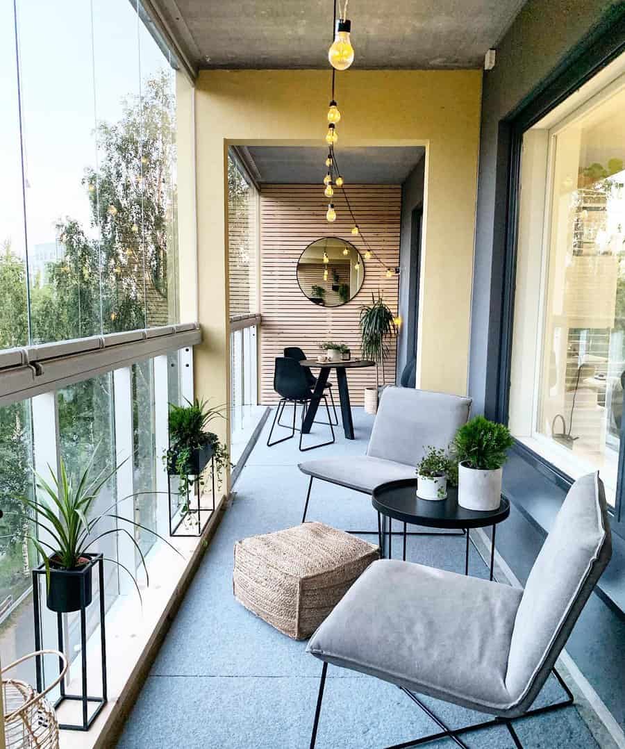 Modern enclosed balcony with sleek gray chairs, a black round table, and hanging string lights. A mirror on the wooden accent wall reflects the cozy ambiance, complemented by potted plants and a woven ottoman.