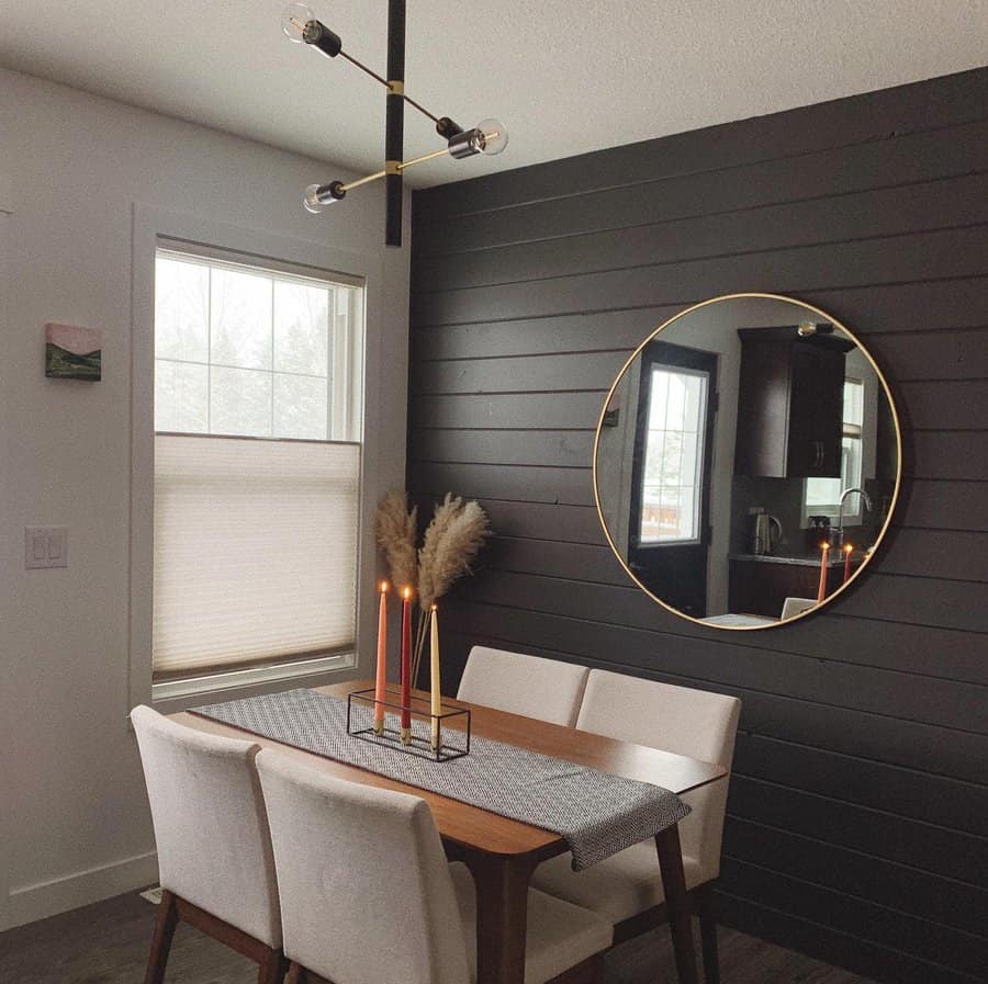 Dining area with a wooden table, four white chairs, candles, and pampas grass; black shiplap wall with a round mirror above the table