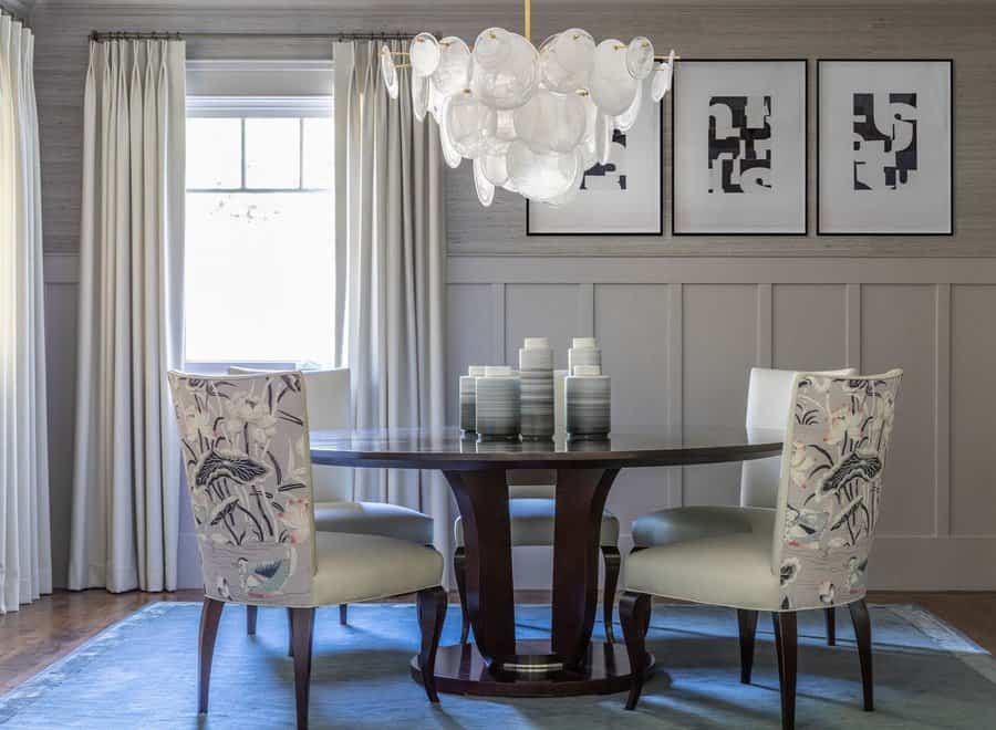 Elegant dining room with a round table, four chairs, modern chandelier, abstract wall art, gray wainscoting, and a light blue rug