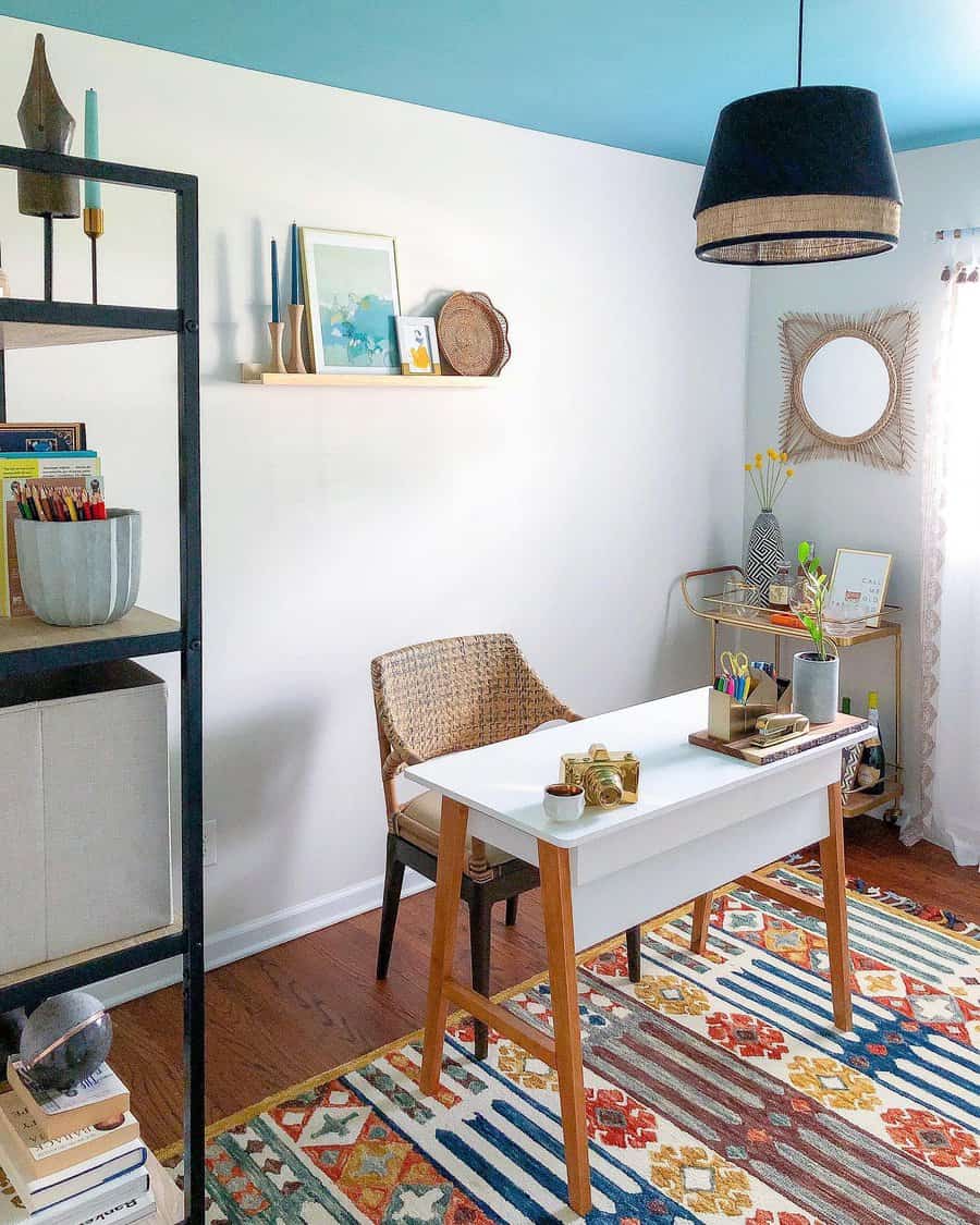 Bright home office with a white desk, wicker chair, colorful rug, shelf, and hanging lamp; decor includes plants and framed art