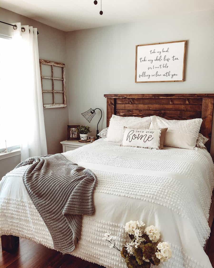 Cozy bedroom with a wooden bed, white bedding, a gray throw, decorative pillows, wall art, and a potted plant by the window