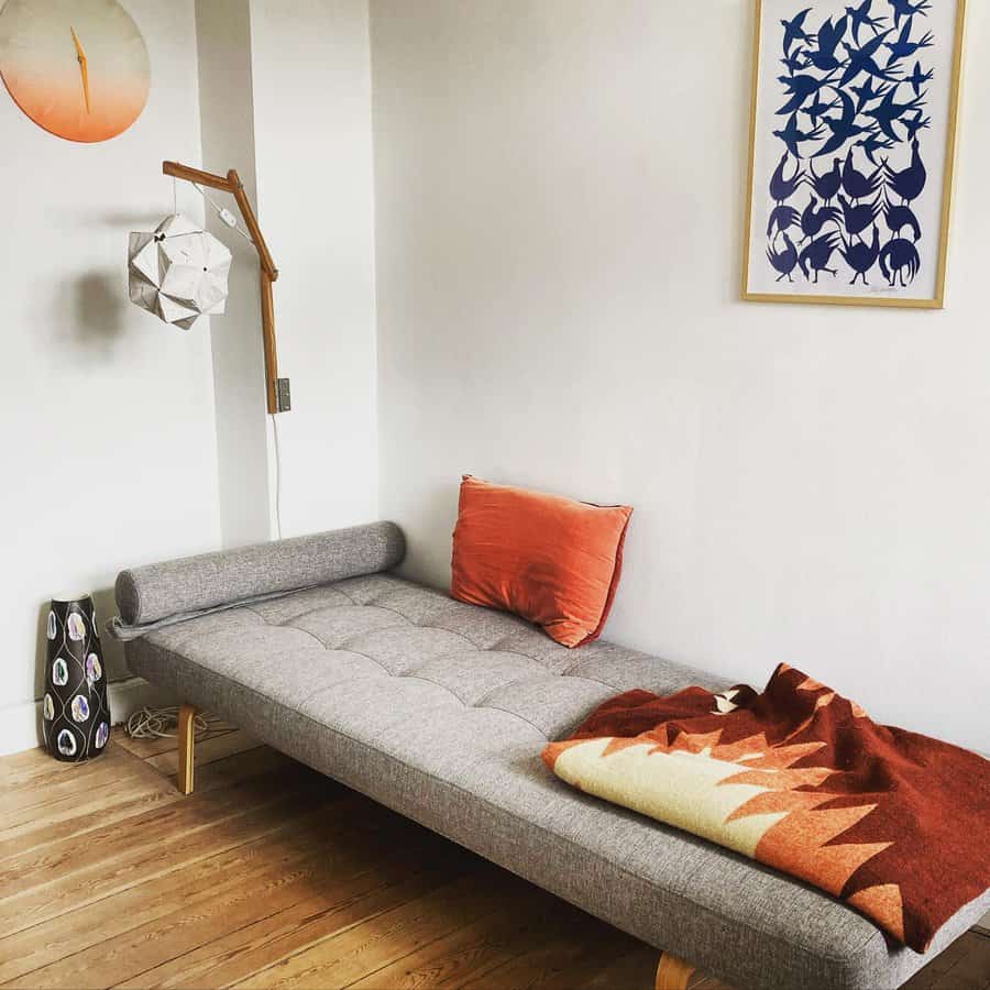 Minimalist room with a gray daybed, orange cushion, geometric artwork, and a cozy blanket on wooden floor