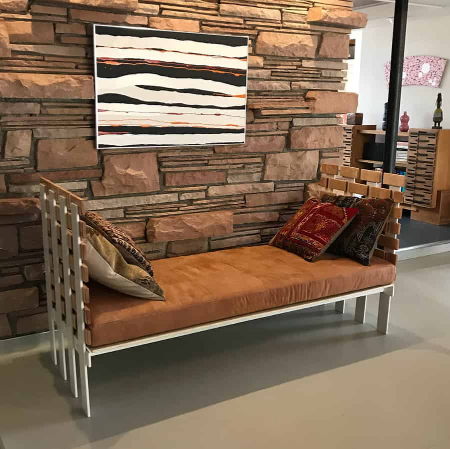 Modern daybed with a sleek white frame, tan suede cushion, and woven side panels, set against a rustic stone wall with bold artwork