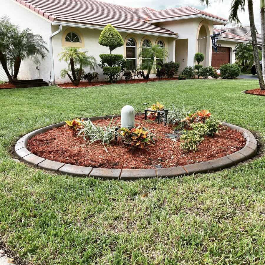 Front yard landscaping with a circular mulch bed, vibrant tropical plants, stone edging, and a neatly maintained lawn