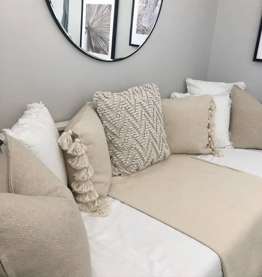 Beige and white daybed with textured pillows, tassels, and a soft throw; round mirror and framed art on the wall; cozy and minimalist