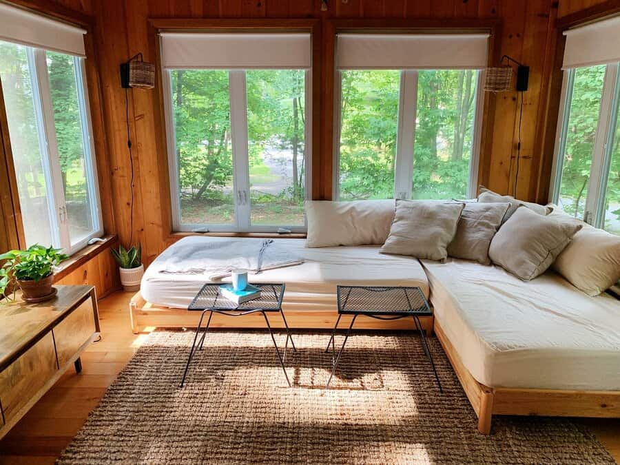 Sunlit room with large windows, L-shaped daybed with pillows, small table, potted plants, and a woven rug on wooden floor