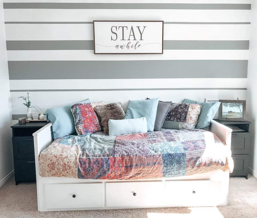 A cozy daybed with a colorful patchwork quilt, surrounded by pillows, framed by striped wallpaper and a "Stay Awhile" sign above