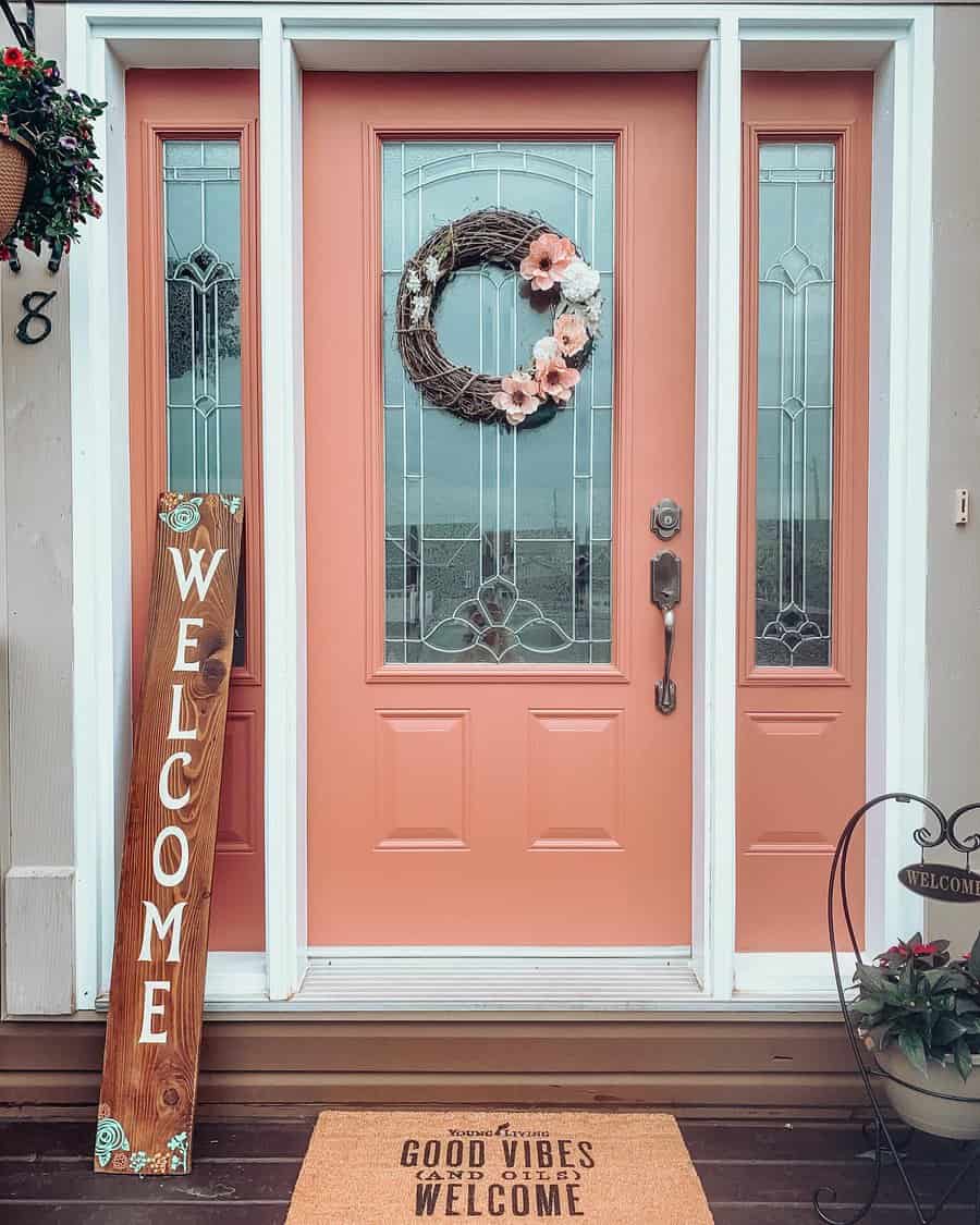 Pink front door with floral wreath, flanked by glass panels; wooden "Welcome" sign and doormat saying "Good Vibes Welcome"