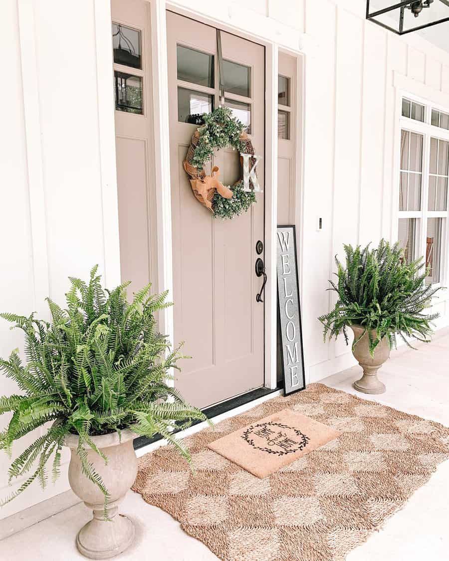 Front porch with beige door, green wreath, "Welcome" sign, two potted ferns, and a woven doormat with a decorative design