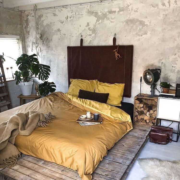 Bedroom with a rustic wooden floor, brown and mustard bedding, plants, a retro lamp, and logs stacked beside a white nightstand