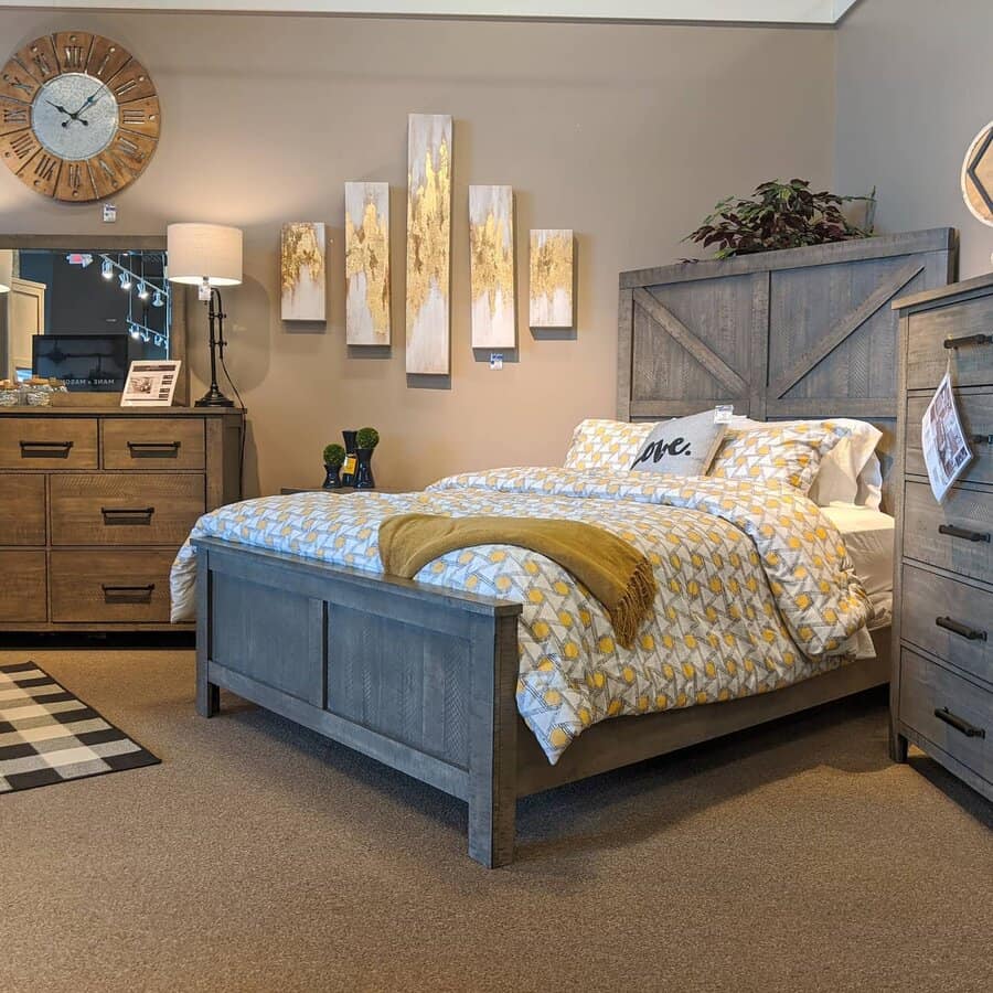 Cozy bedroom with a gray wood bed, yellow-patterned bedding, artwork above, dresser, lamp, and plants; warm and inviting decor