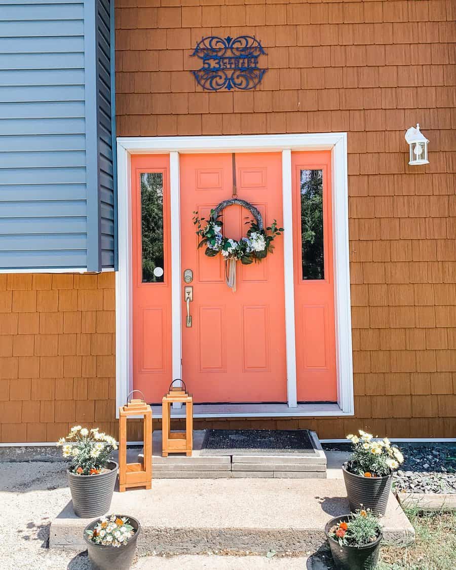 A coral-colored door with a floral wreath, flanked by lanterns and potted flowers, against a brown shingle exterior