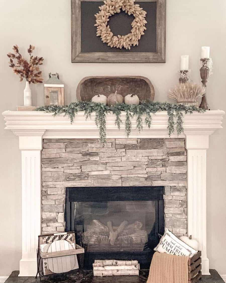 A stone fireplace with autumn decor: a wreath, candles, pumpkins, greenery, books, and a cozy basket with blankets