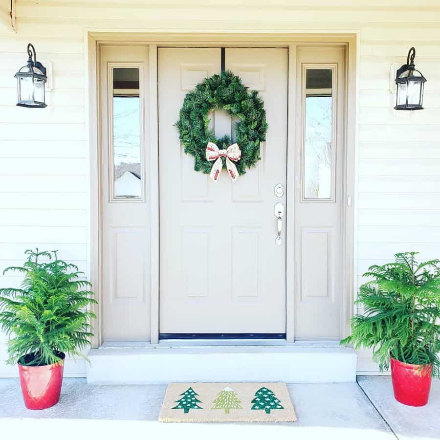 Front door with a green wreath, two small trees in red pots, and a doormat featuring green Christmas trees, flanked by two lanterns