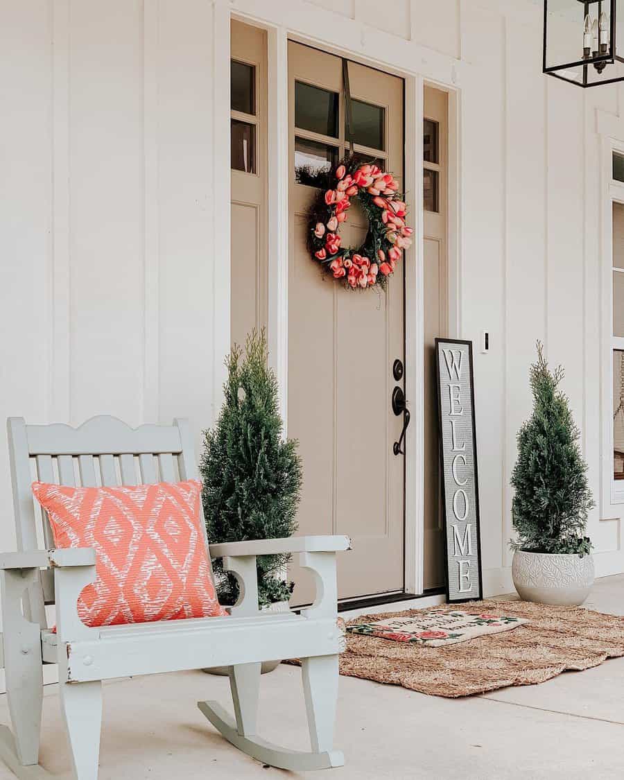 Front porch with a rocking chair and orange cushion, two potted plants, tulip wreath on a beige door, and a "Welcome" sign beside a mat