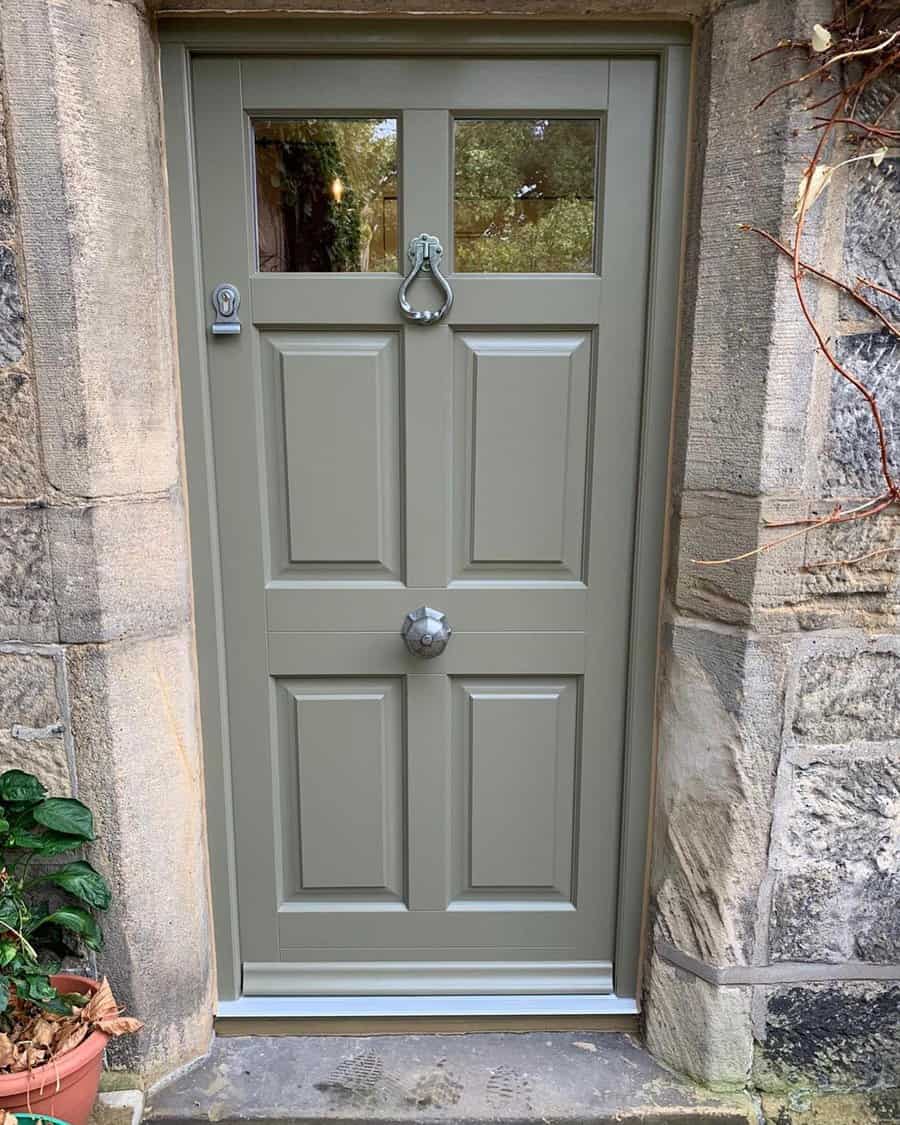 A sage green wooden door with two knockers, surrounded by stone walls and a potted plant on the left