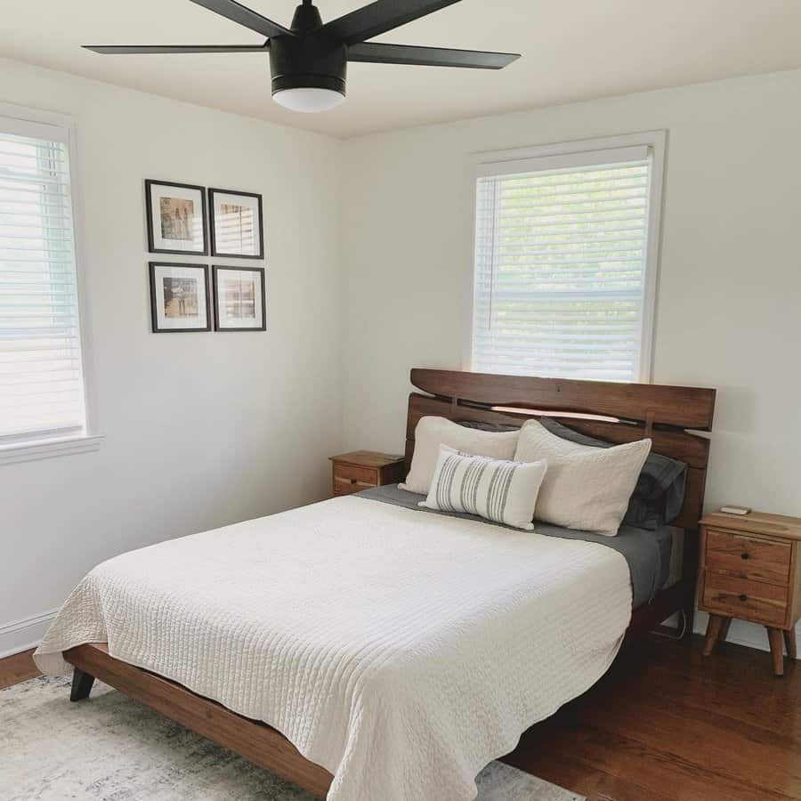 Minimalist bedroom with a wooden bed frame, white bedding, and two bedside tables; a black ceiling fan above and framed pictures on the wall