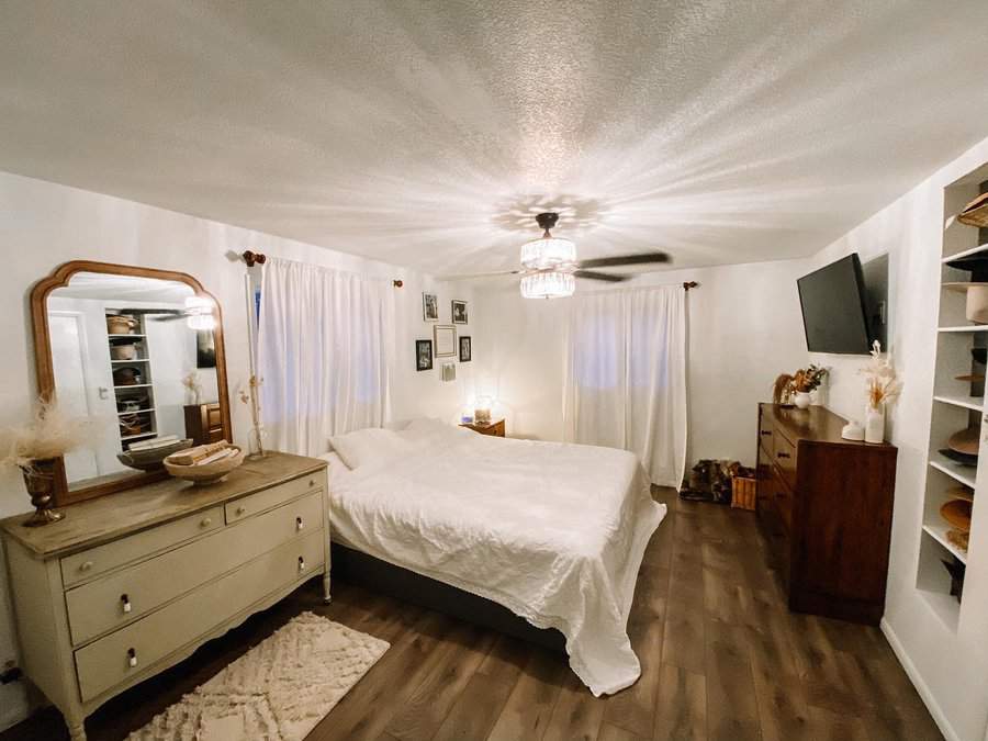 Master bedroom with a bed, dresser, and wall-mounted TV; elegant chandelier lighting, white curtains, and wooden floor enhance the warmth