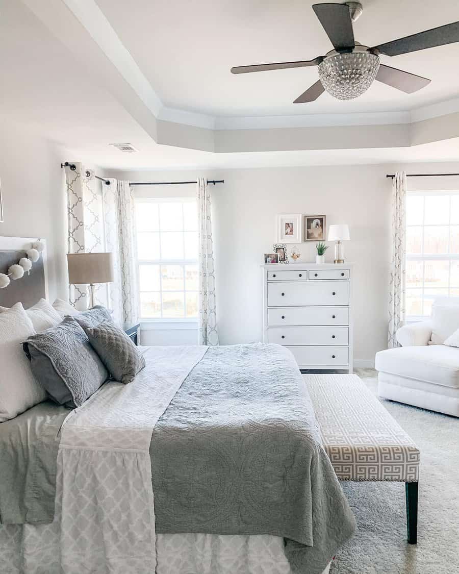 Bright bedroom with a large bed, gray and white linens, white dresser, armchair, and two windows with patterned curtains, ceiling fan above