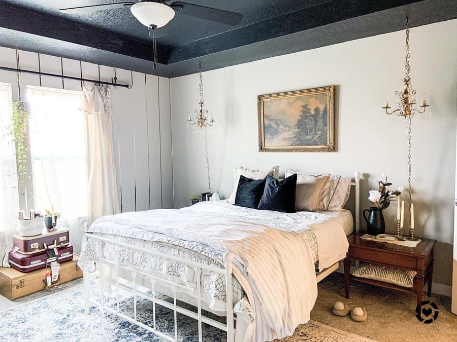 A master bedroom with a white bed, black ceiling fan, chandeliers, and vintage decor, including stacked suitcases and a landscape painting