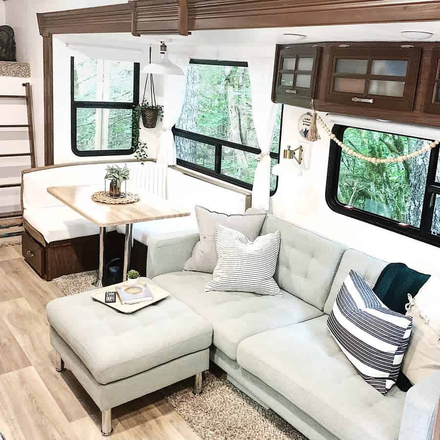 A neutral-toned RV living area featuring a cozy sectional sofa with striped pillows, a wooden dining table, and large windows offering a peaceful, nature-filled view.