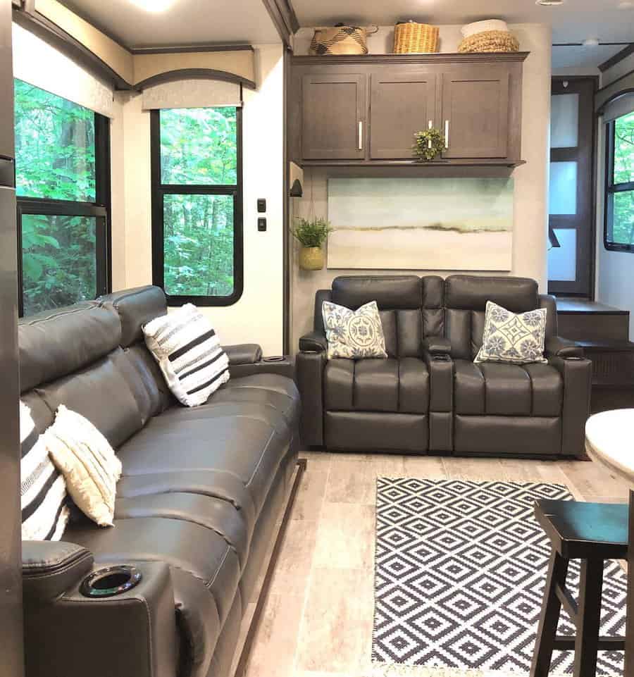 A sleek, neutral-toned RV living room with black leather furniture, patterned pillows, a cozy rug, and an inviting view through large windows.