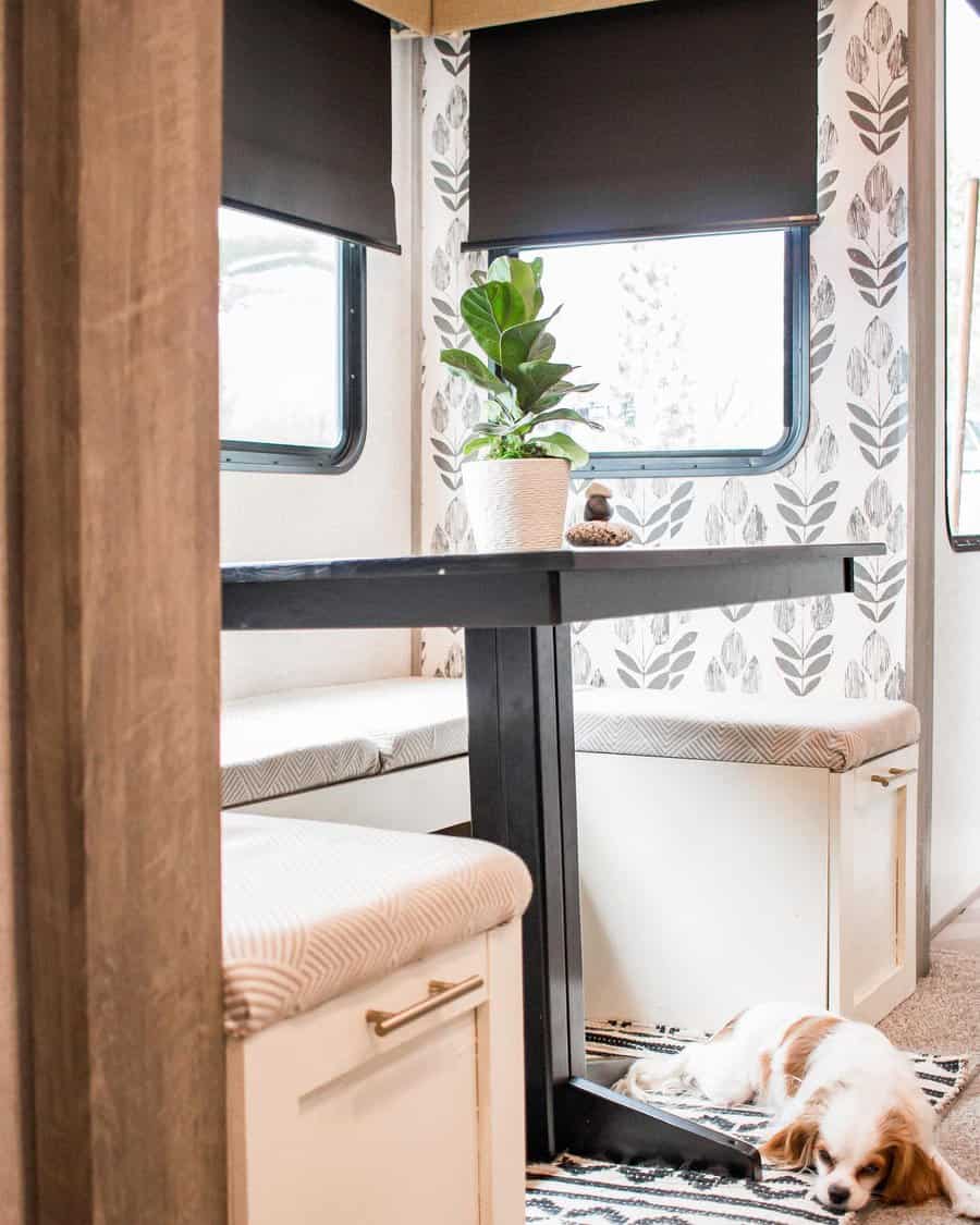 A cozy, neutral-toned RV dining area with a simple table, cushioned seats, a leafy plant, and a cute dog resting on the floor, creating a welcoming and peaceful space.