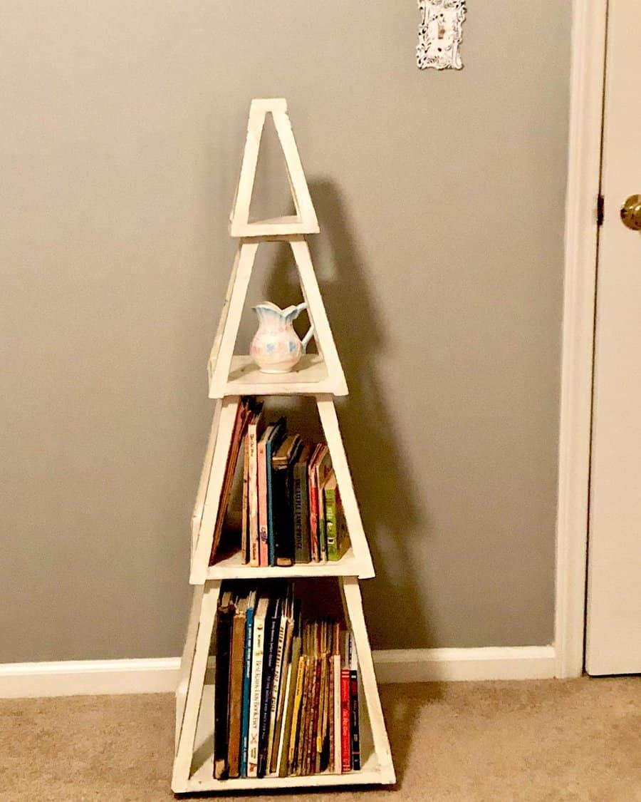 Rustic wooden pyramid-shaped bookshelf holding children's books and a decorative pitcher, placed against a neutral-toned wall.