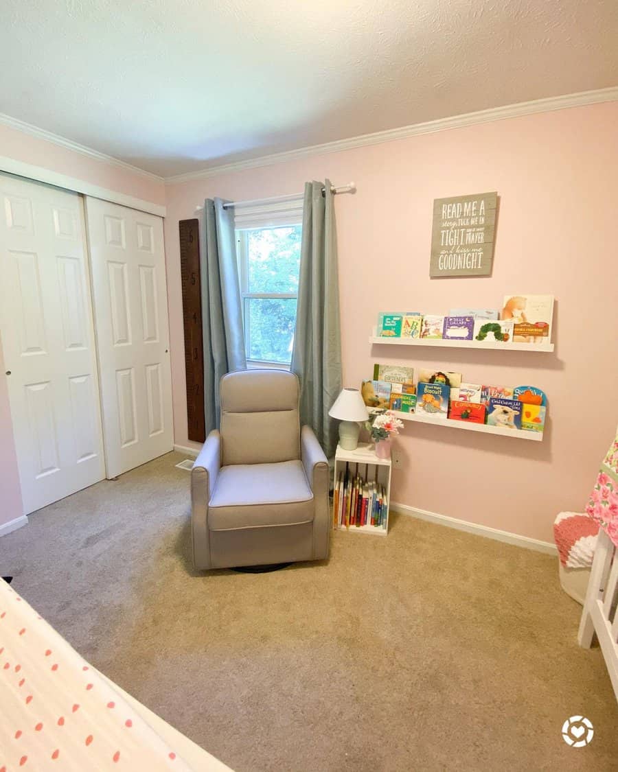 Soft pink nursery with a cozy reading nook, featuring floating bookshelves, a comfy gray rocking chair, and a small bookshelf beside a lamp.