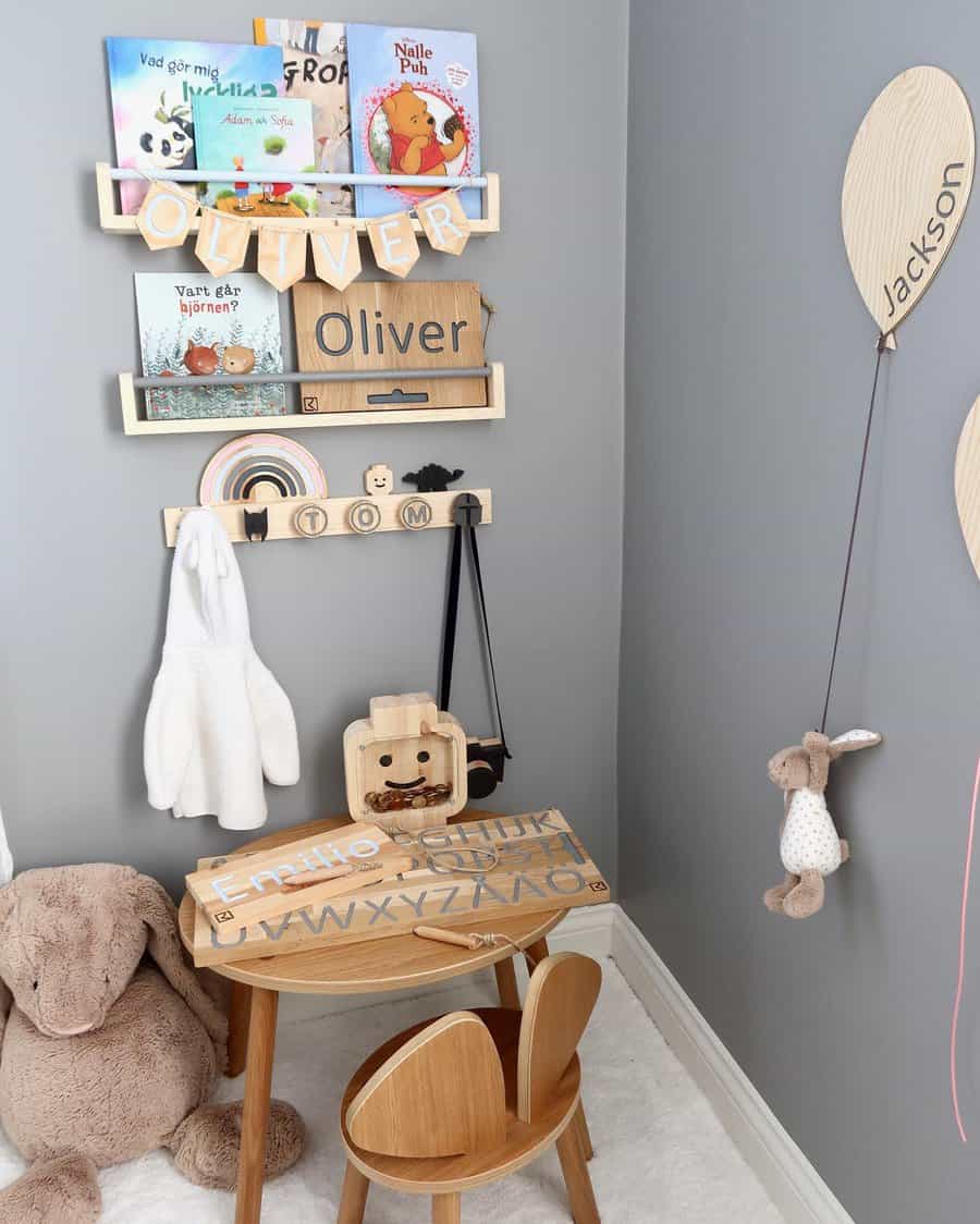 Adorable nursery reading corner with wooden wall-mounted bookshelves, a small wooden table and chair, and plush toys against a gray wall.