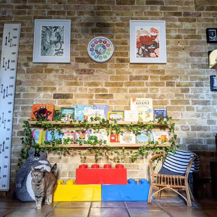 Cozy reading nook with wall-mounted bookshelves decorated with greenery, large LEGO storage blocks, a rattan chair, and a cat on a brick wall backdrop.