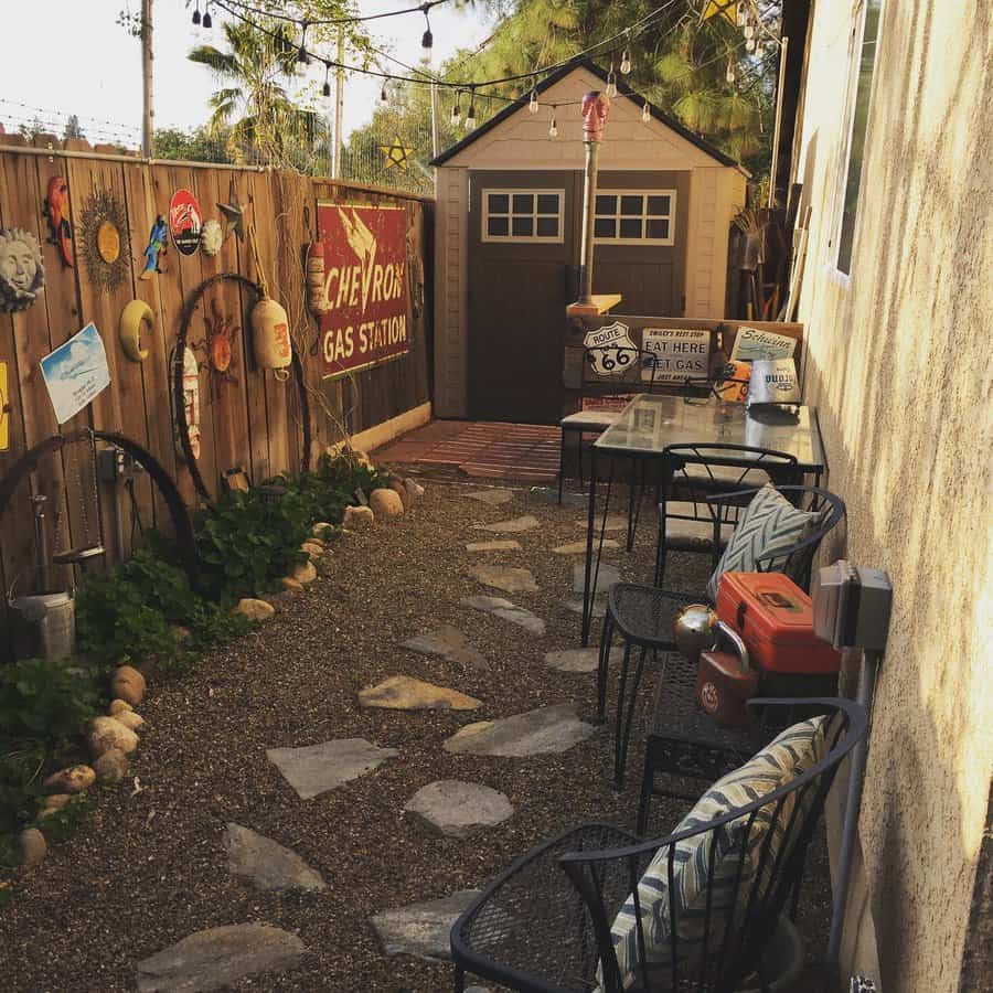 Narrow backyard with a stone path, vintage signs, metal chairs, a red cooler, and a small shed at the end under string lights