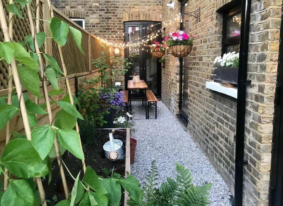 Narrow garden courtyard with string lights, a wooden table, hanging flower baskets, and a brick wall. Pathway is covered with gravel