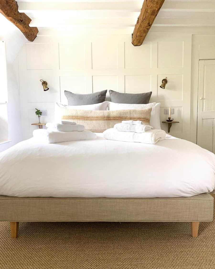 White bedroom with a large bed, white linens, folded towels, pillows, wooden beams on the ceiling, and wall-mounted lamps