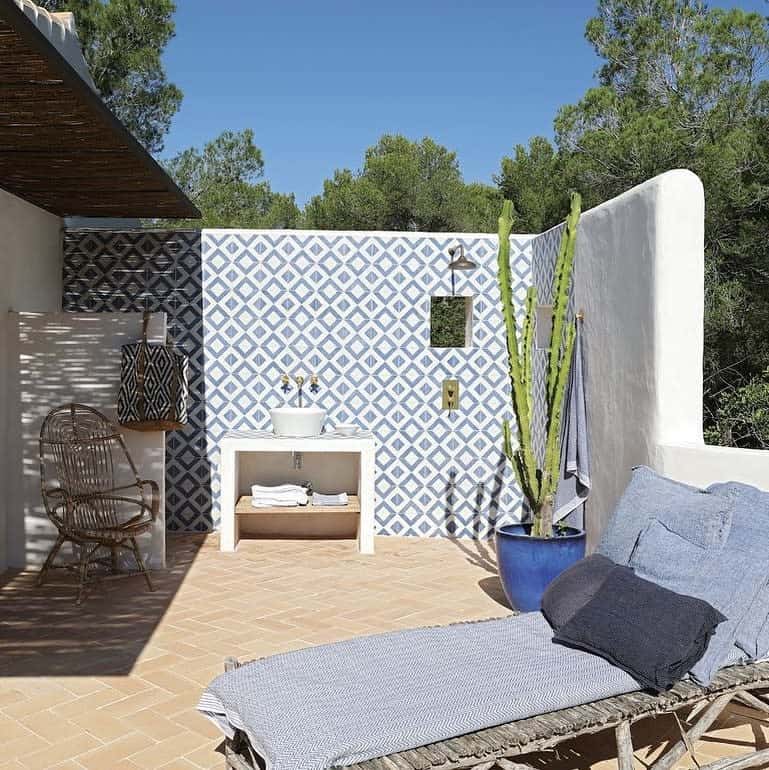 Outdoor patio with a blue patterned tile wall, sink, potted cactus, wicker chair, and a cushioned bench; trees in the background