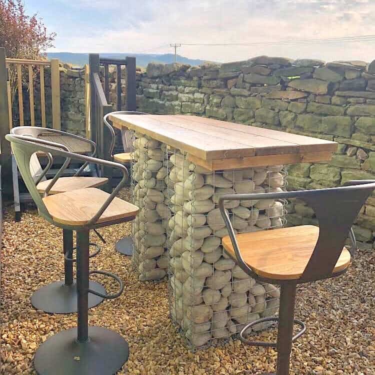 Outdoor patio with a tall wooden table, stone base, and four matching chairs on a gravel surface, surrounded by a stone wall