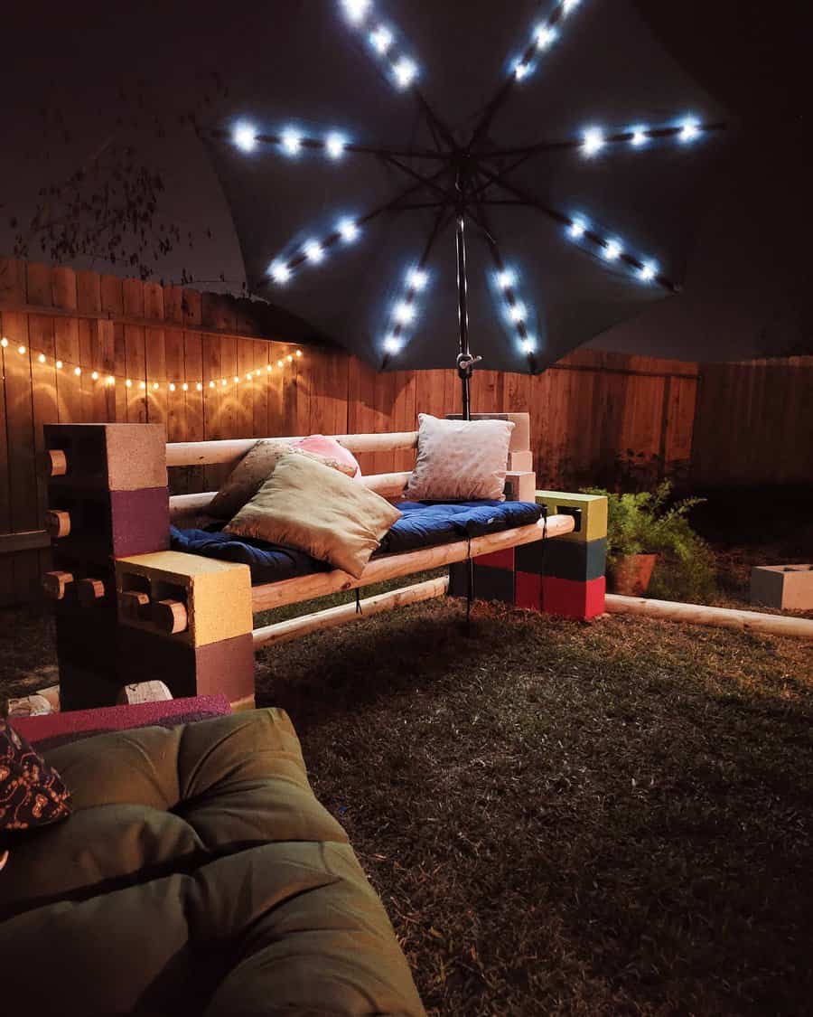 Cozy DIY cinder block bench with colorful blocks, soft cushions, and string lights, set under an illuminated umbrella for a charming backyard retreat