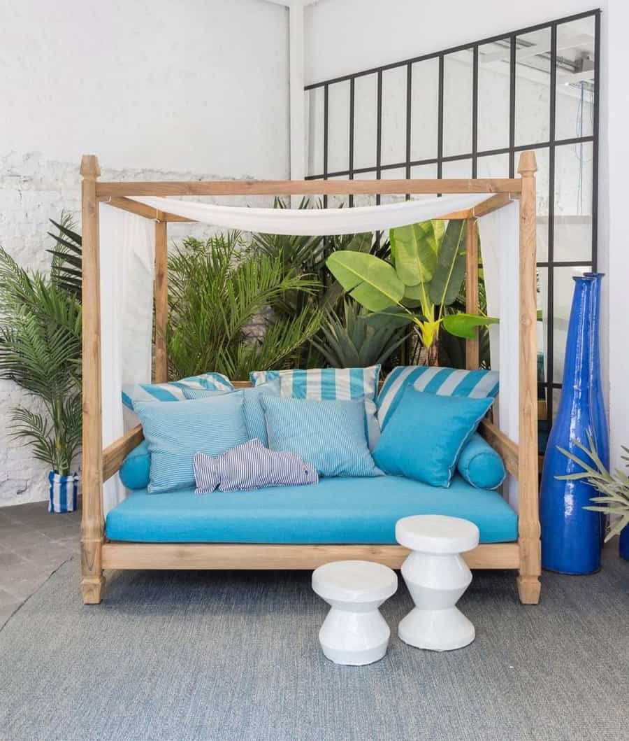 Wooden canopy daybed with blue cushions and tropical plants, white stools in front, against white walls and a large window backdrop