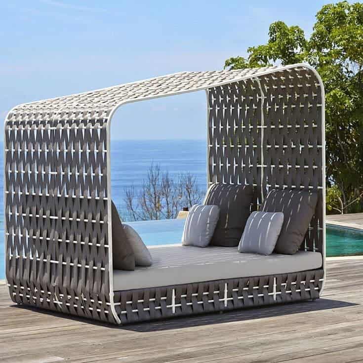 Woven outdoor daybed with cushions under a canopy, set on a wooden deck overlooking the ocean and trees