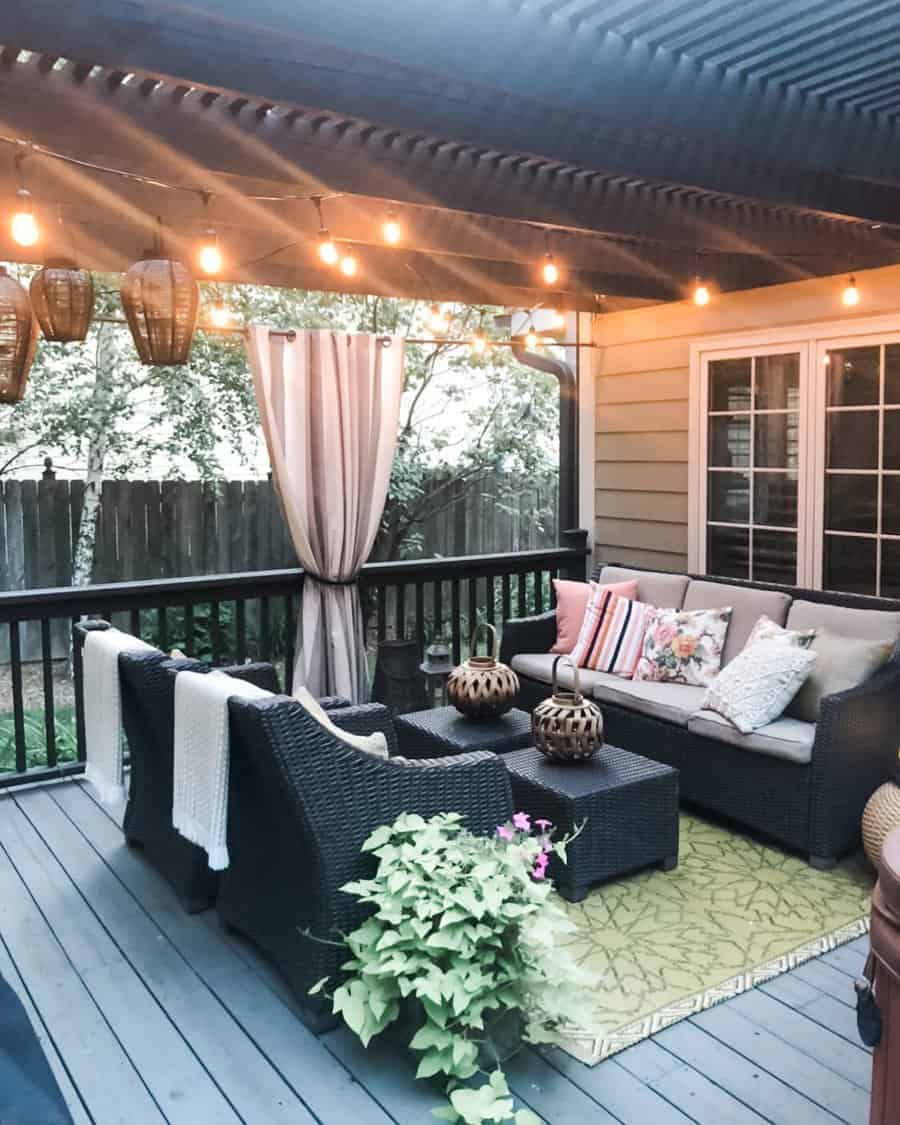 A cozy outdoor deck with wicker furniture, cushions, a rug, and hanging string lights. Potted plants add greenery to the space.