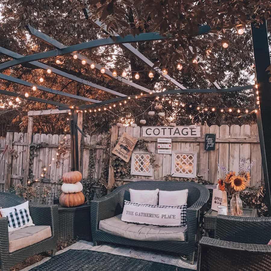 Cozy outdoor patio with wicker seating, string lights, pumpkin decor, and rustic signs; sunflowers in vase on table