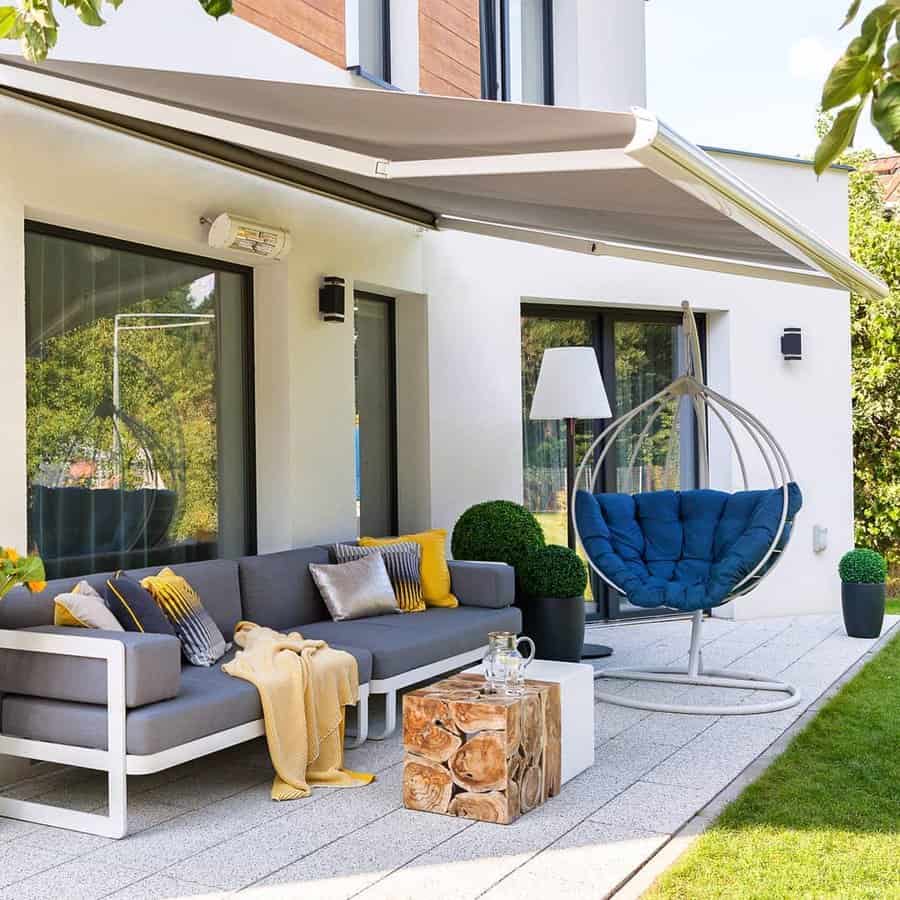 Modern patio with gray sofa, yellow and gray pillows, a hanging chair, wood stump table, and retractable awning