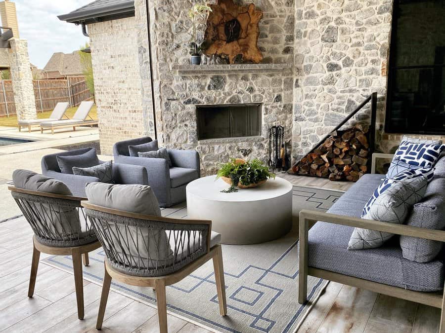 Outdoor patio with wicker chairs and a sofa around a round coffee table, stone wall with a fireplace and logs stacked beside it