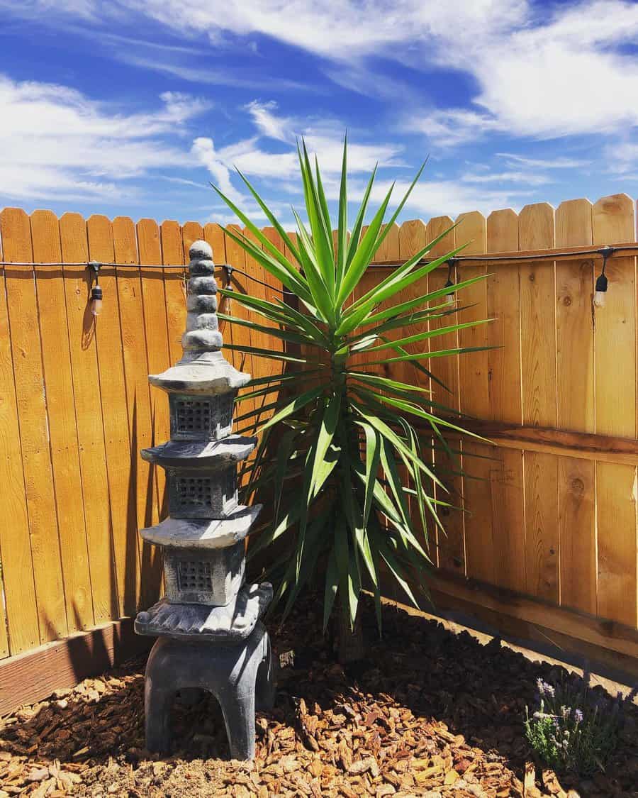 A small pagoda statue and a tropical plant in a garden corner, bordered by a wooden fence under a blue sky with clouds